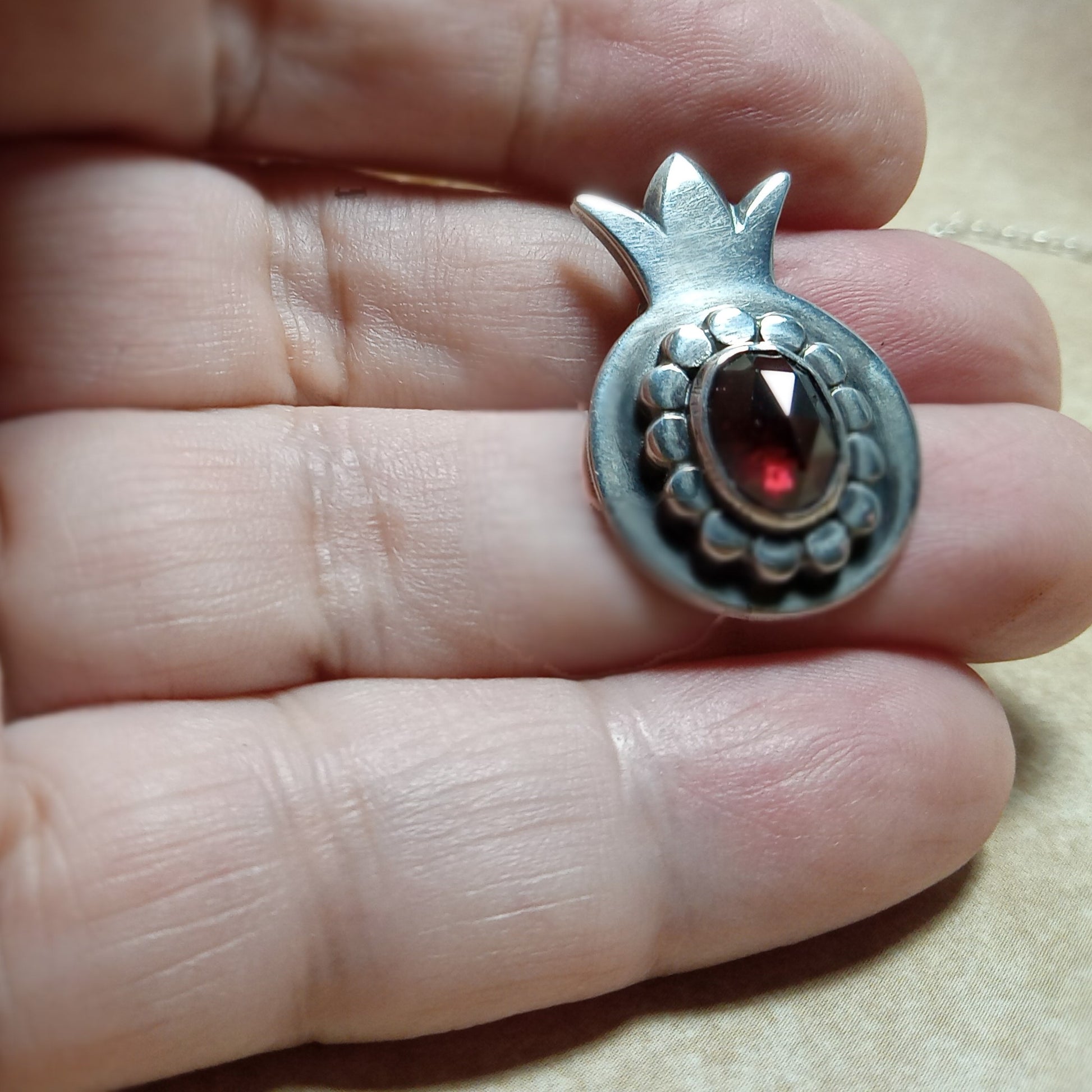 Silver pomegranate-shaped pendant with a red garnet gemstone held between fingers.
