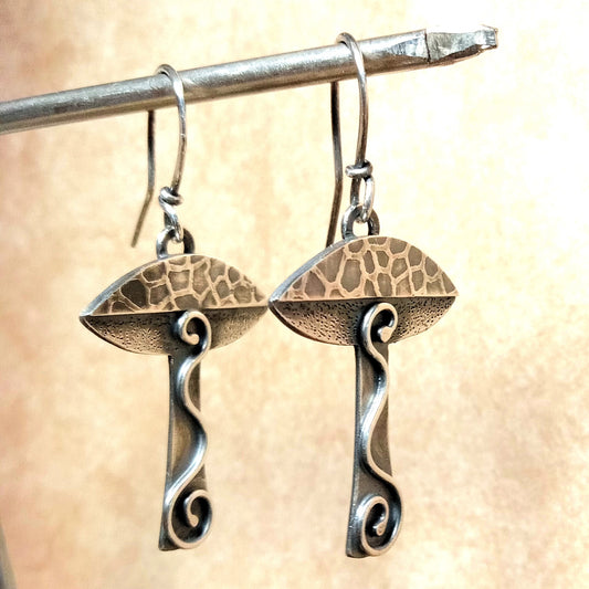 Handcrafted Argentium sterling silver mushroom earrings with hammered caps and swirling textured stems, oxidized and polished for a rustic, mystical woodland aesthetic. Suspended from metal prop.