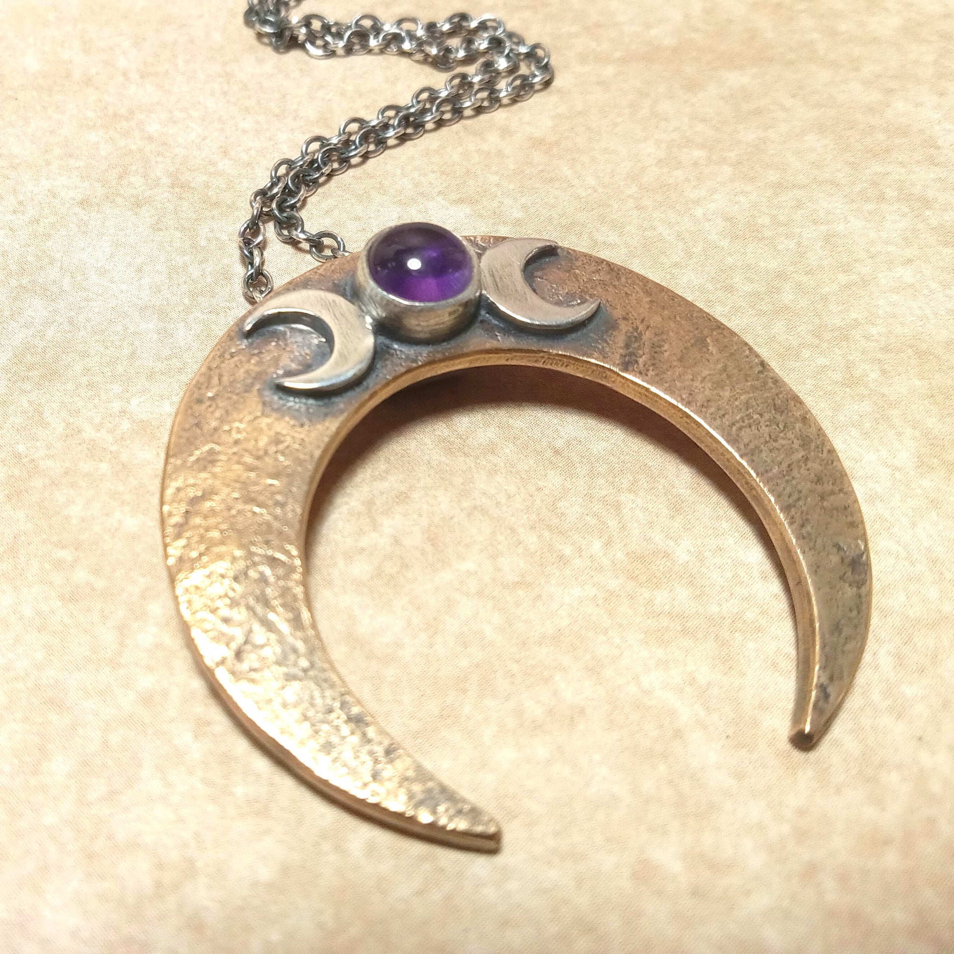 Crescent moon-shaped pendant with two small moons and an amethyst gemstone on a beige background.