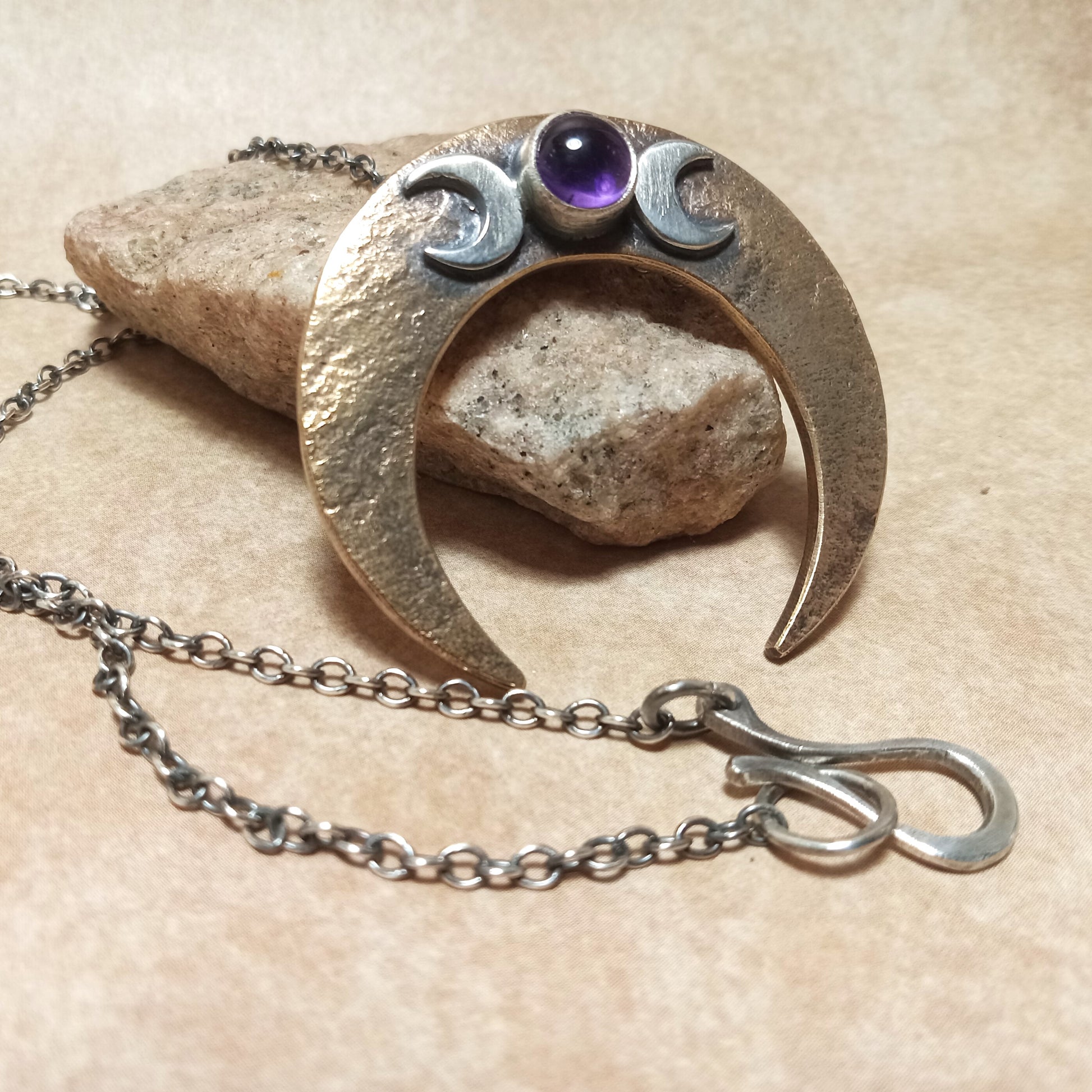Bronze and Silver pagan crescent moon pendant with a amethyst gemstone, showing chain and handmade findings on a beige background.
