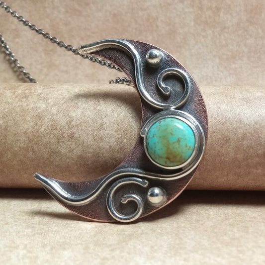 Copper and Silver crescent moon pendant with turquoise stone on a beige background.