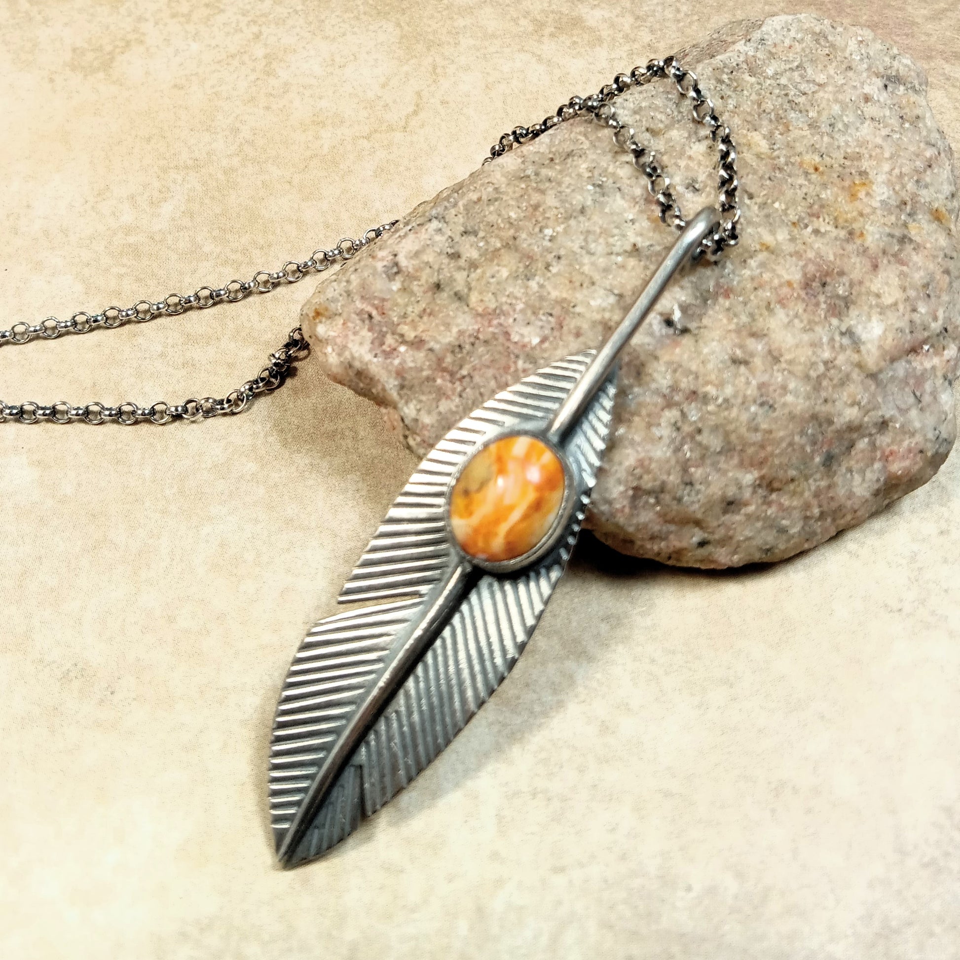 Argentium Silver feather-shaped pendant with an orange spiny oyster cabochon on a SS chain, resting on a rock.