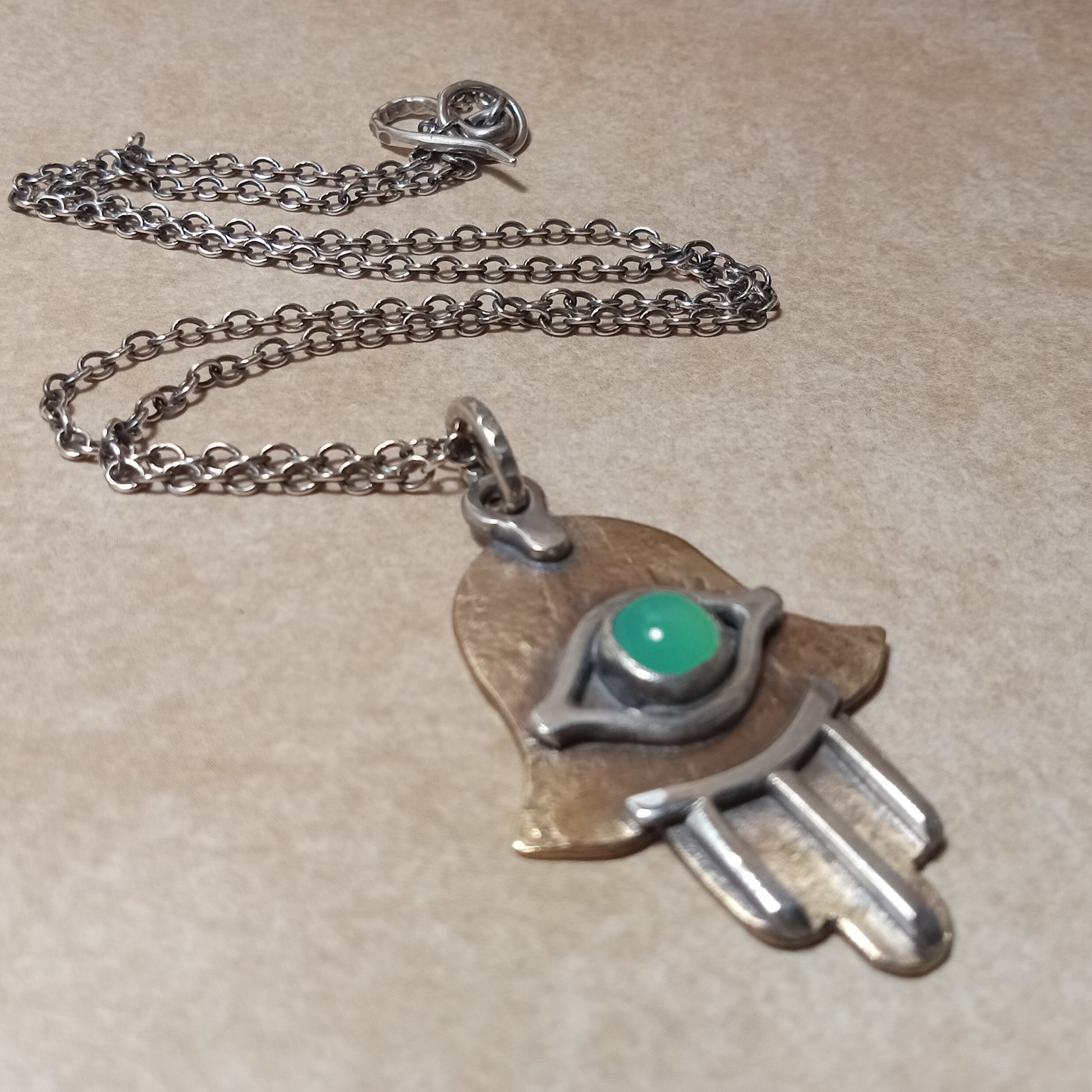 Silver necklace with a hand-shaped pendant featuring a green gemstone on a beige background. SS chain and handmade findings.