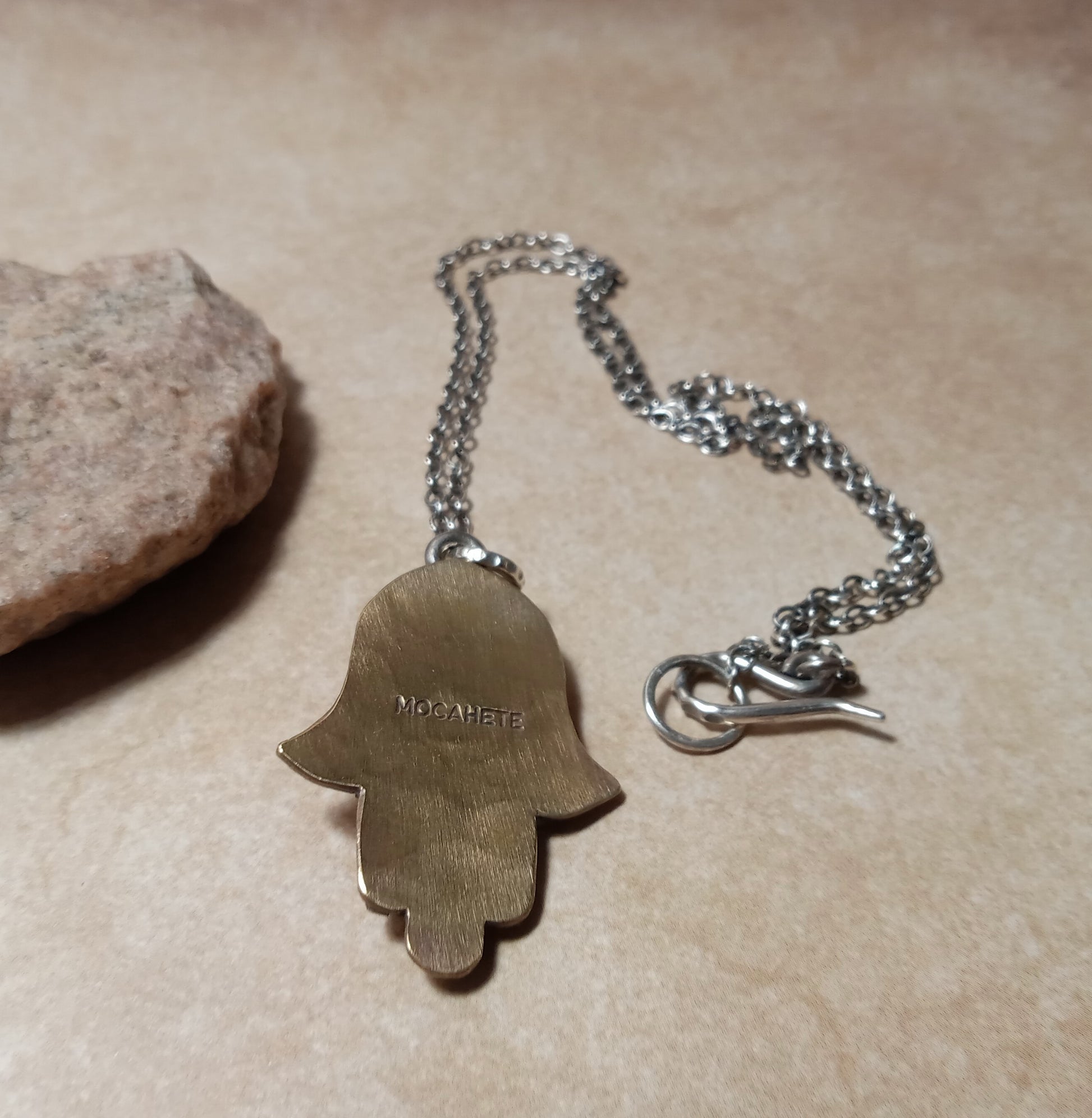 Reverse side of bronze and Silver Hamsa necklace showing makers mark on a beige surface.