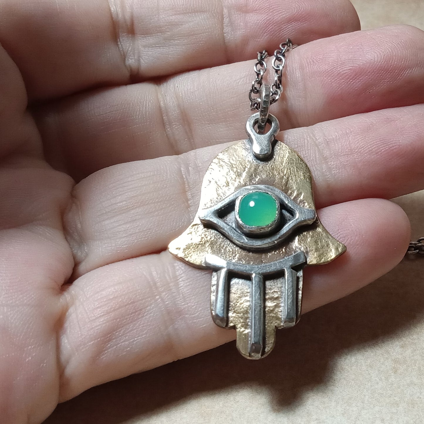 Hand holding a silver and bronze pendant with a green gemstone eye .design.