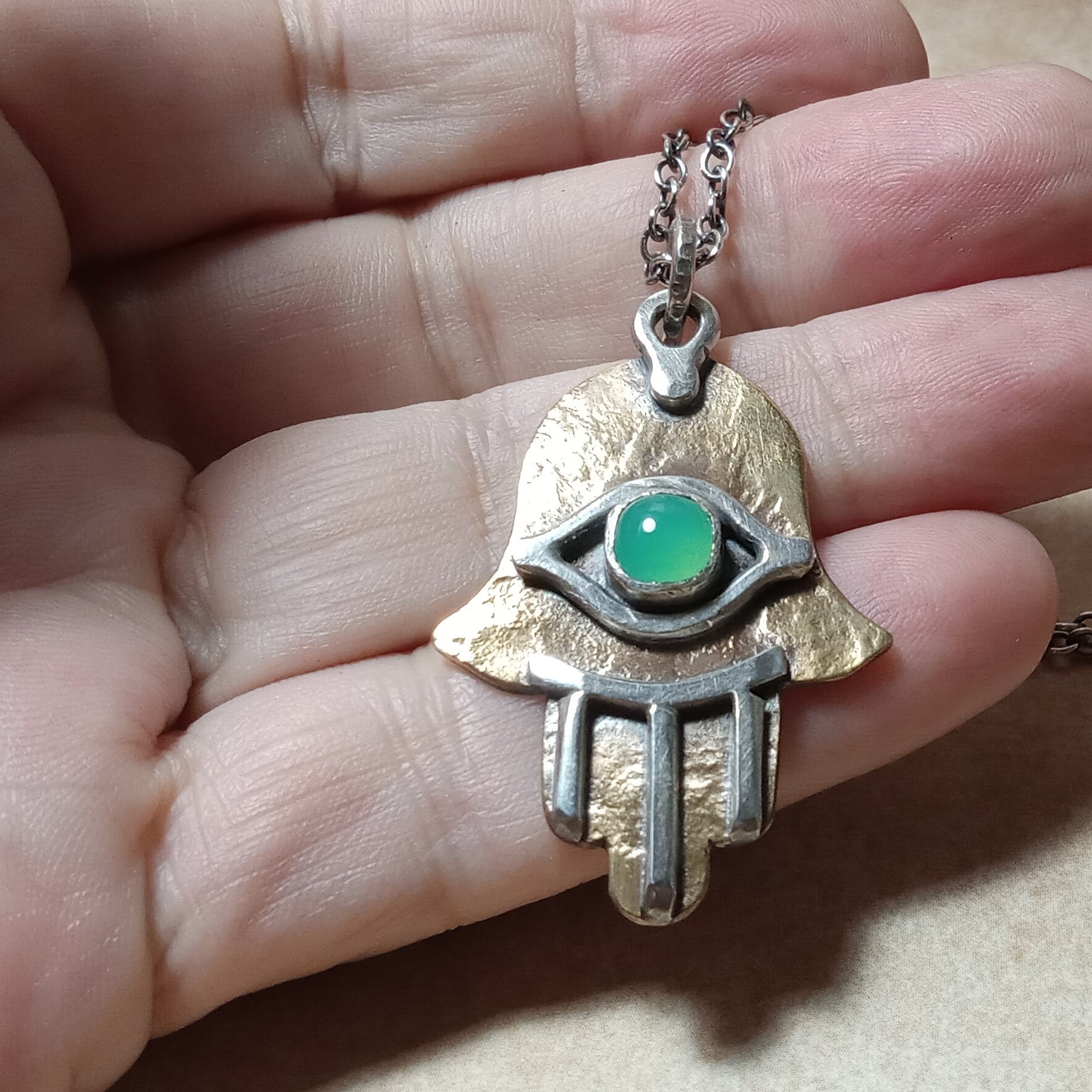 Hand holding a silver and bronze pendant with a green gemstone eye .design.