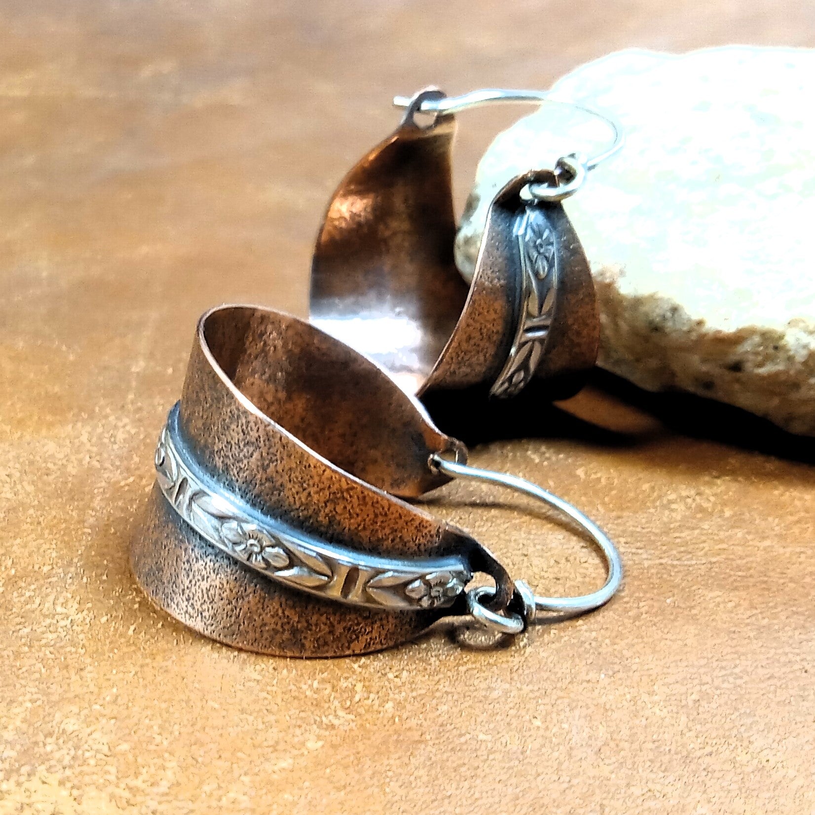 Rustic copper basket-shaped hoop earrings with silver floral detail on brown surface against stone