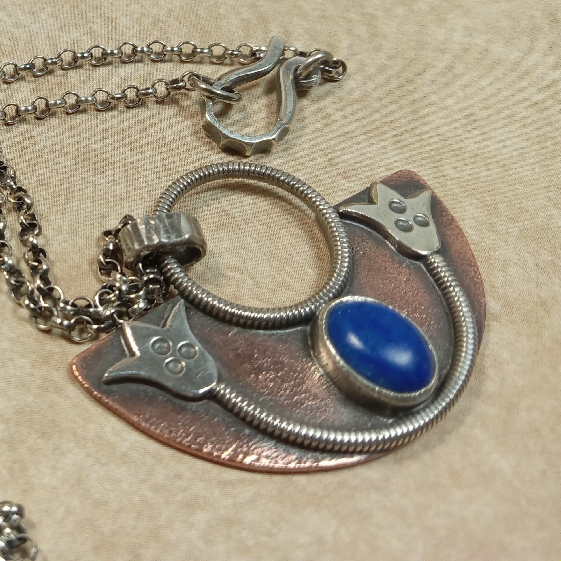 Close-up of handcrafted lotus pendant showing texture, fine silver bezel, clasp and lapis centerpiece on beige background.