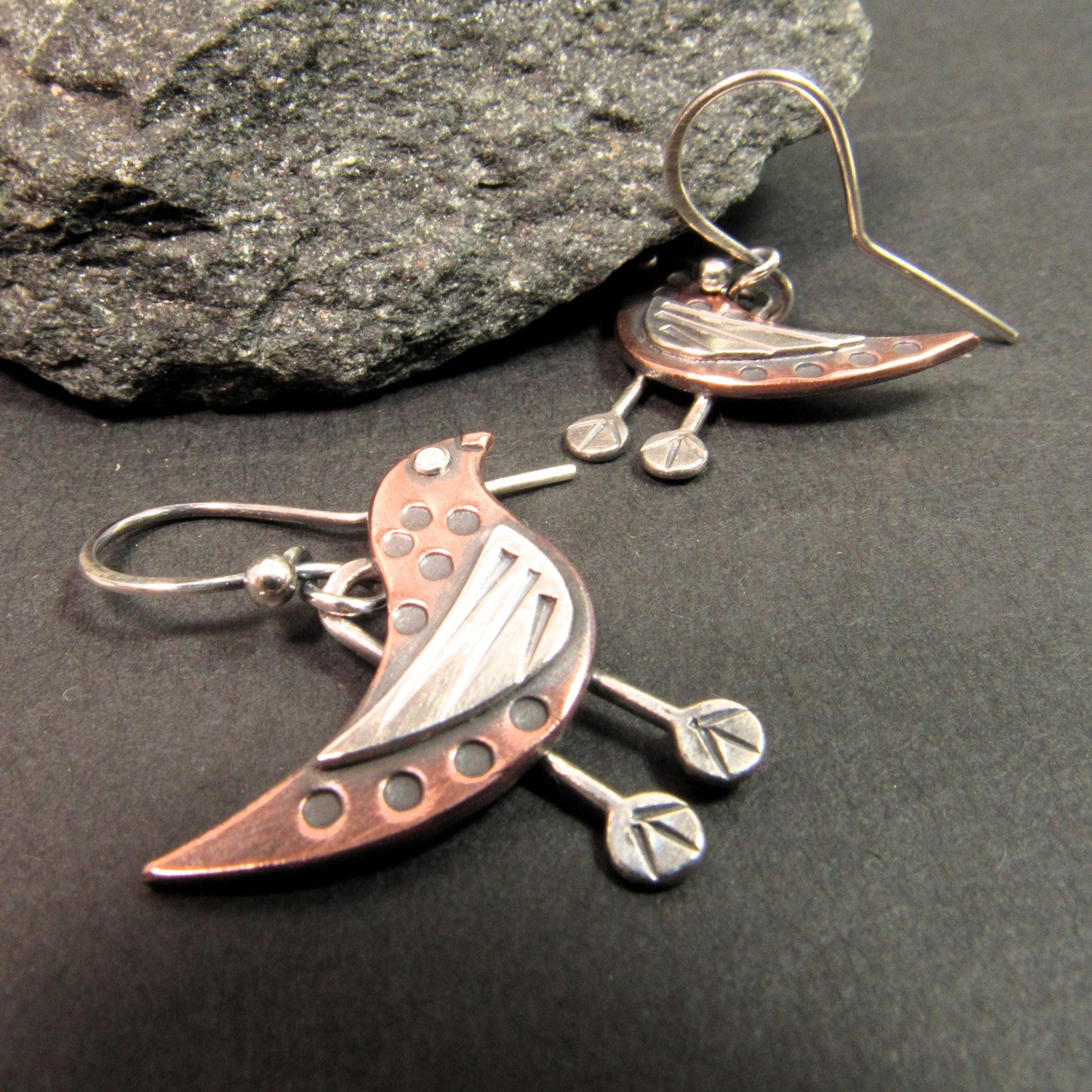 Whimsical handcrafted bird earrings in copper and Argentium silver with riveted detailing, artisan made by Mocahete. On dark background with rock.