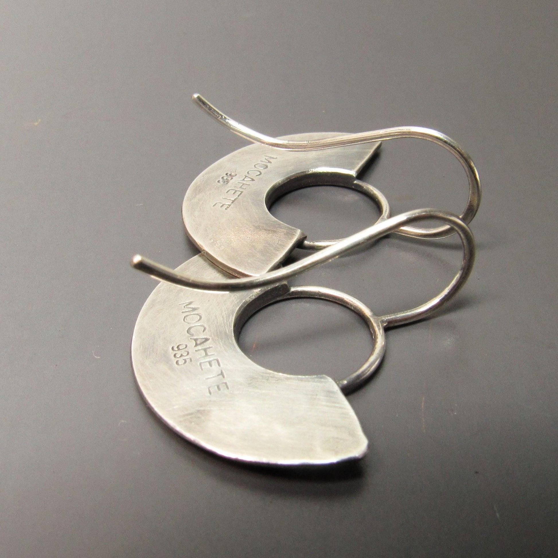Backside of handmade silver fan earrings with makers mark on a gray background.