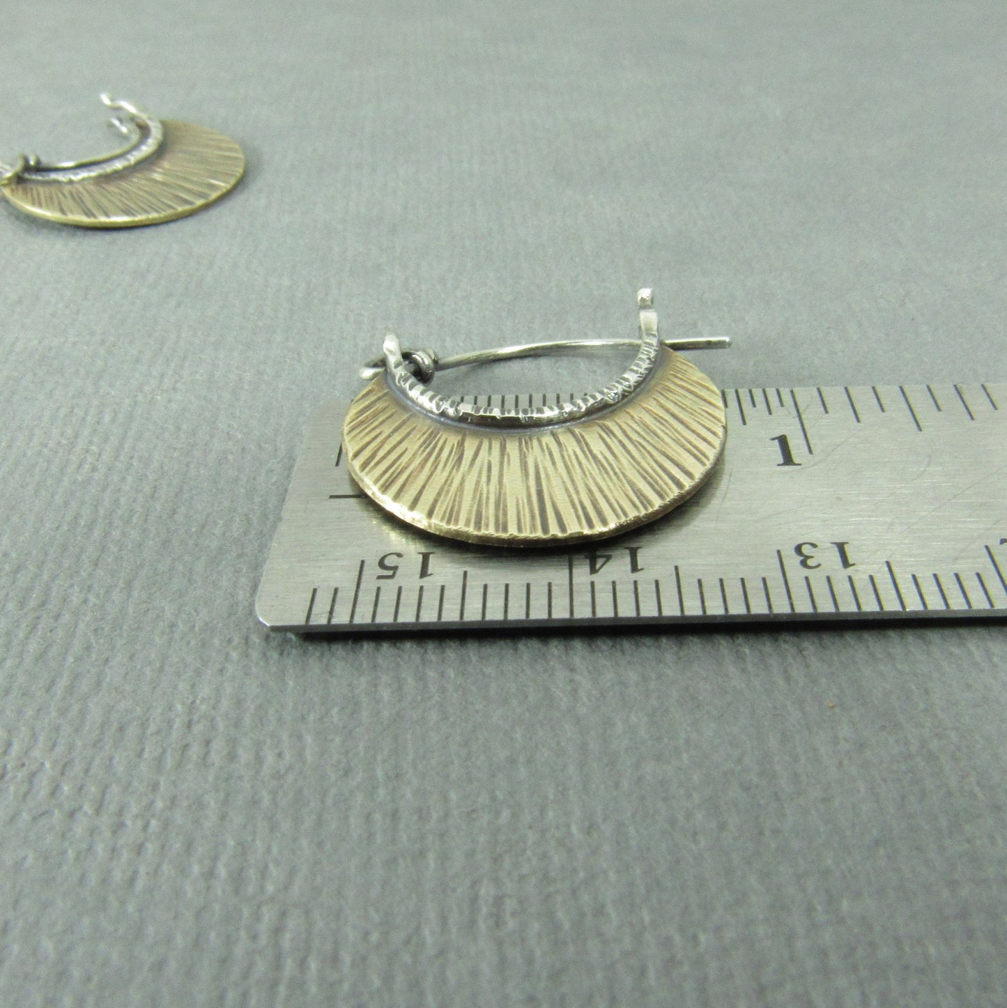 Bronze and silver hoop earrings by Mocahete on a ruler for scale on a gray background.