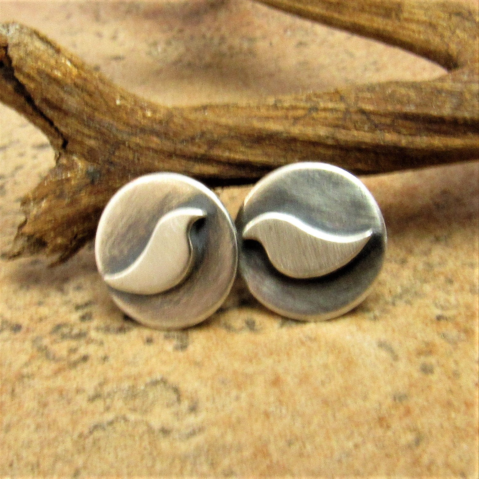 Small sterling silver bird stud earrings with oxidized details on round disks, handcrafted by Mocahete. Photographed against driftwood on stone surface.