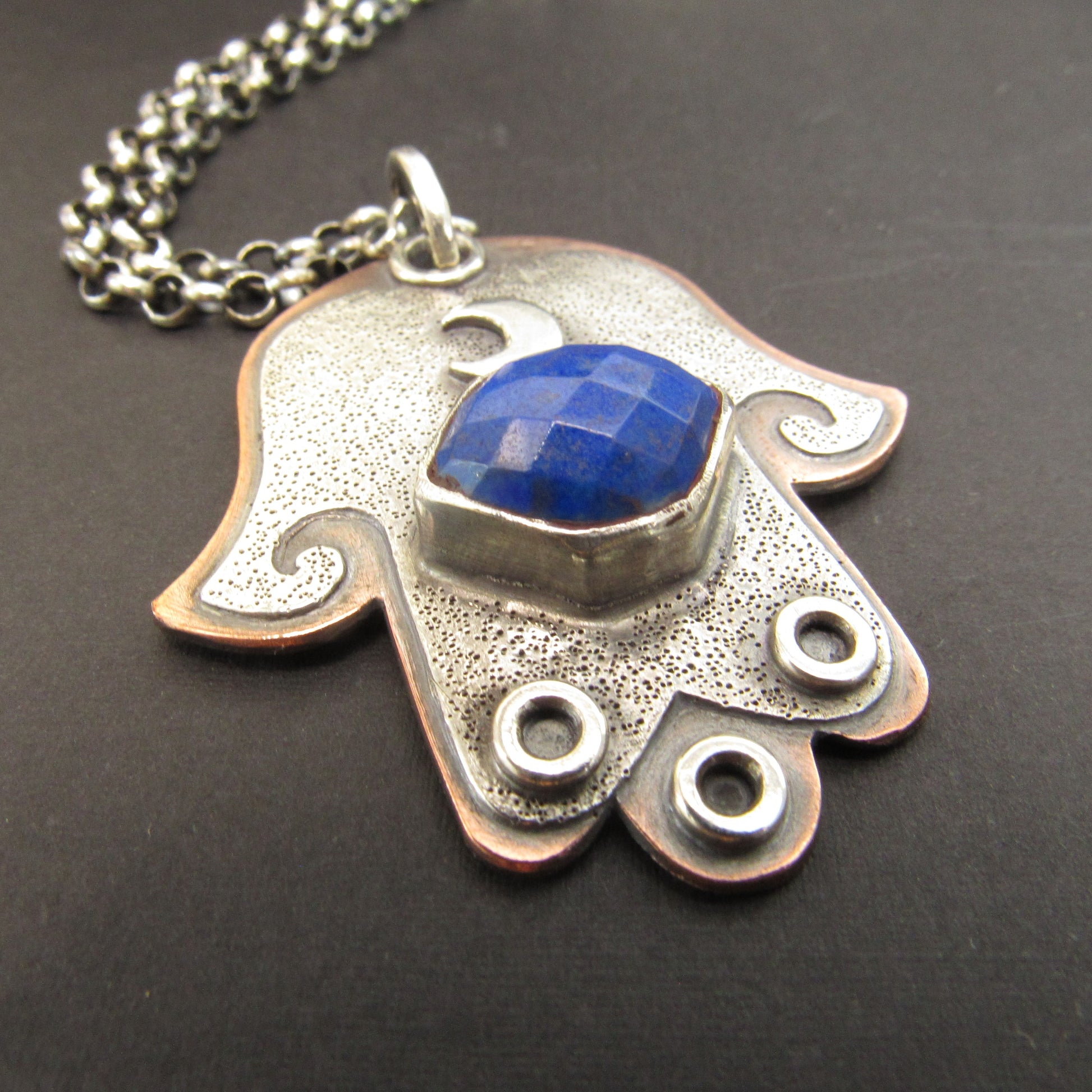 Silver and copper Hamsa pendant with blue stone on a dark background.