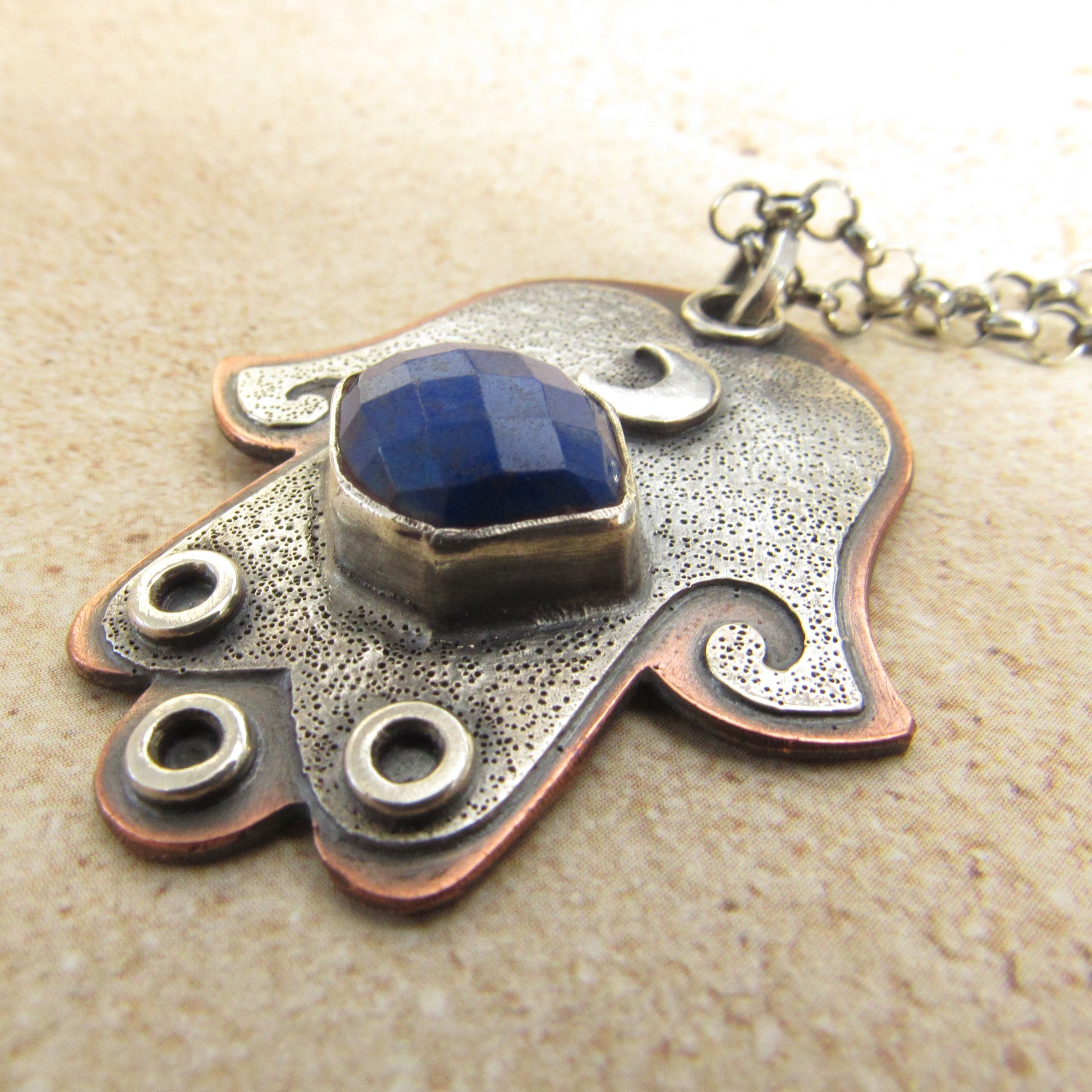Hamsa Jewelry with blue stone and textured metal design on a beige background