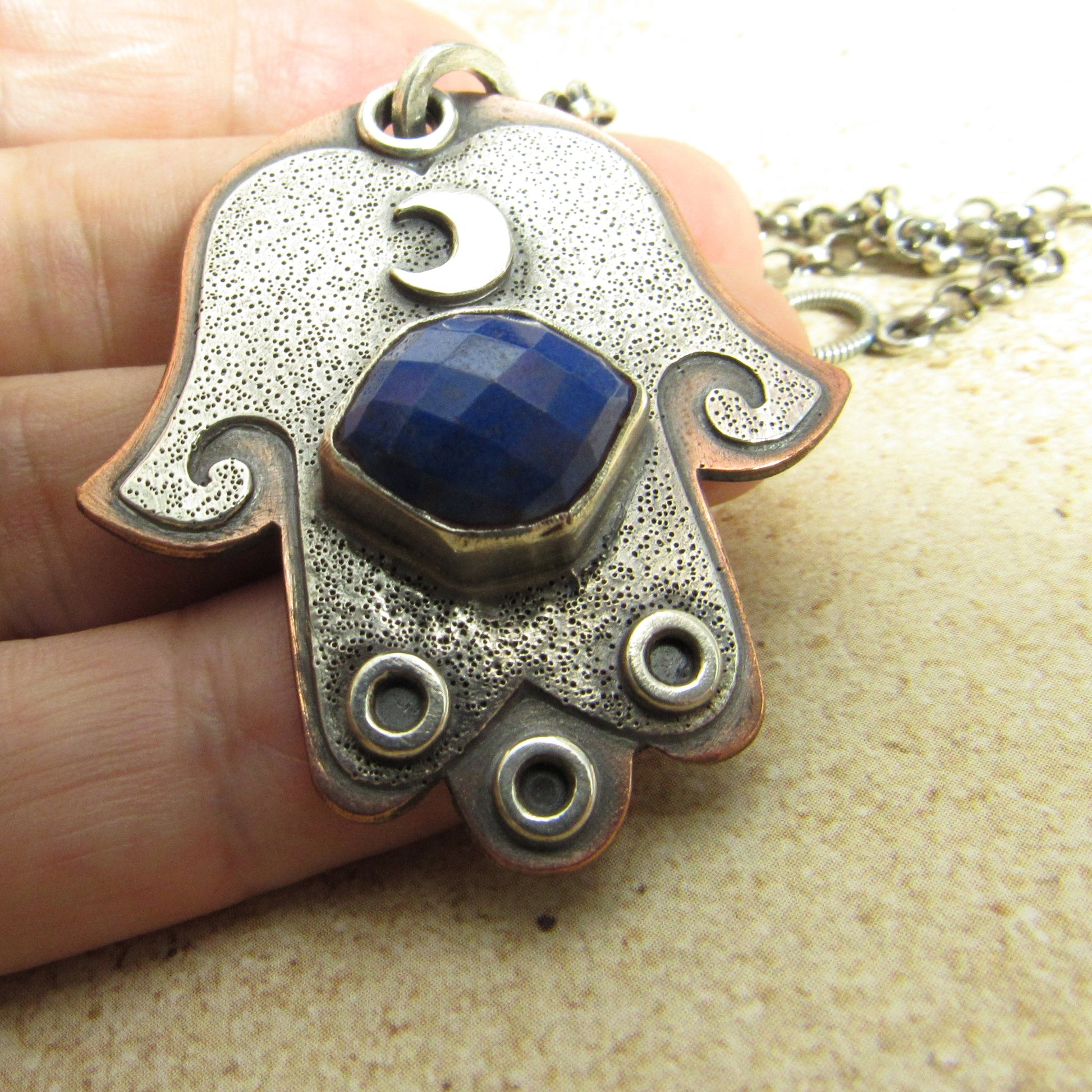 Handmade Hamsa pendant with blue stone and textured surface held by a hand on a beige background