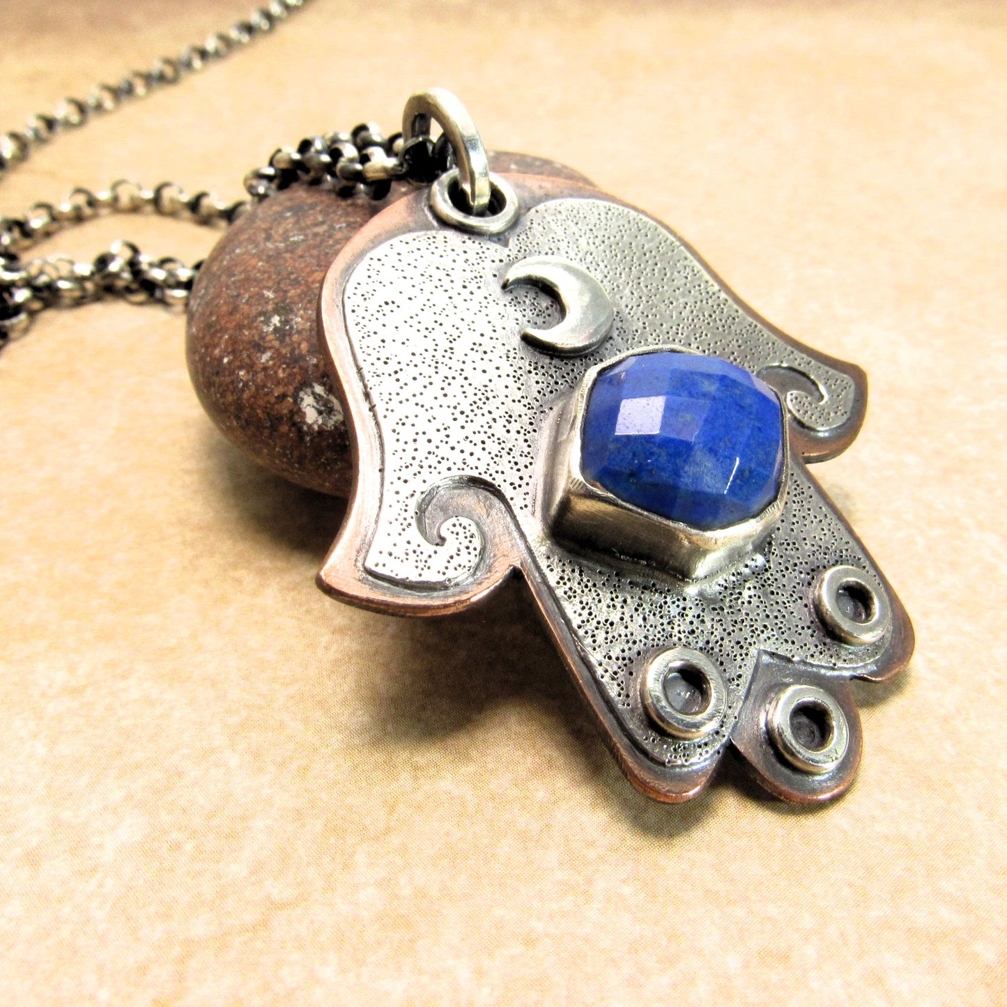 Silver and copper Hamsa hand pendant with a blue lapis gemstone on a beige background with rock