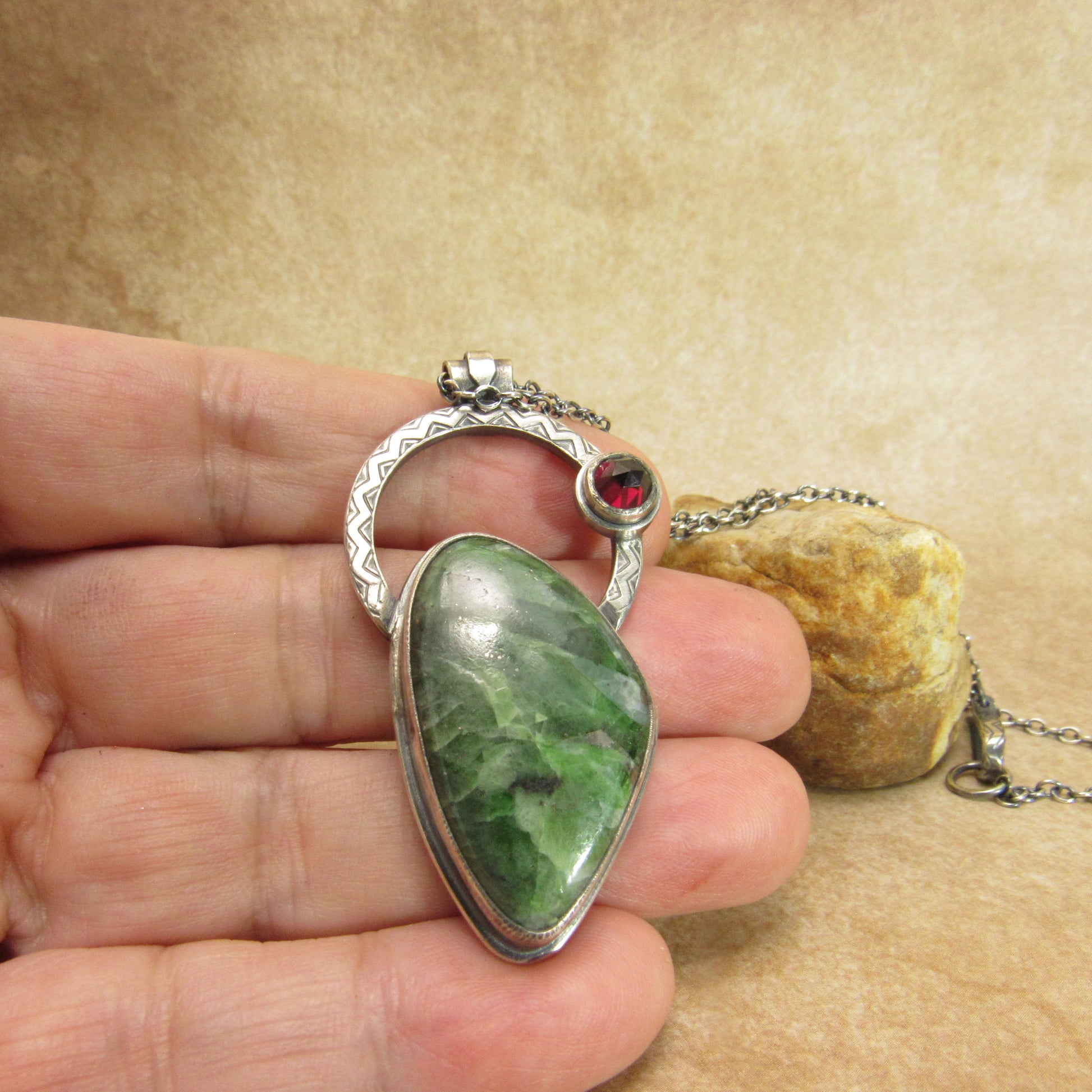 Silver and chrome diopside pendant necklace with red garnet gemstone. Photographed in hand on a neutral sand colored background.