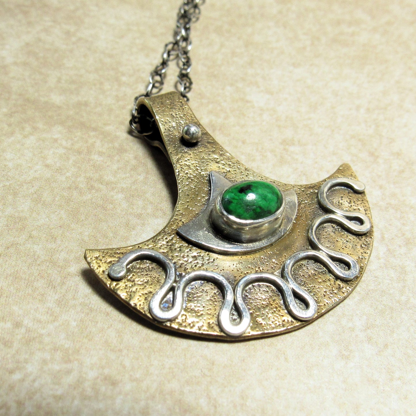 Bronze and silver tribal style pendant with green gemstone on a beige background.