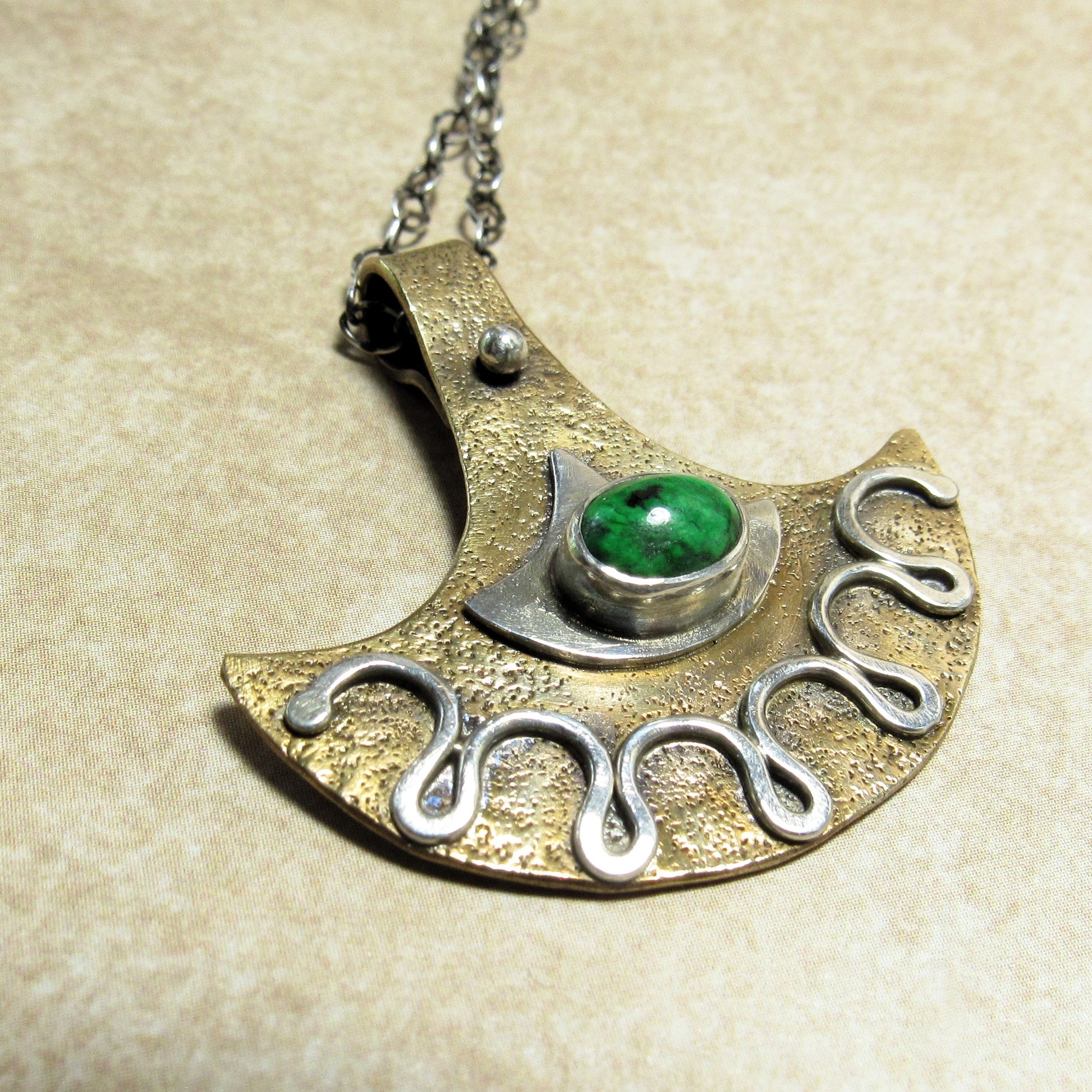 Bronze and silver tribal style pendant with green gemstone on a beige background.