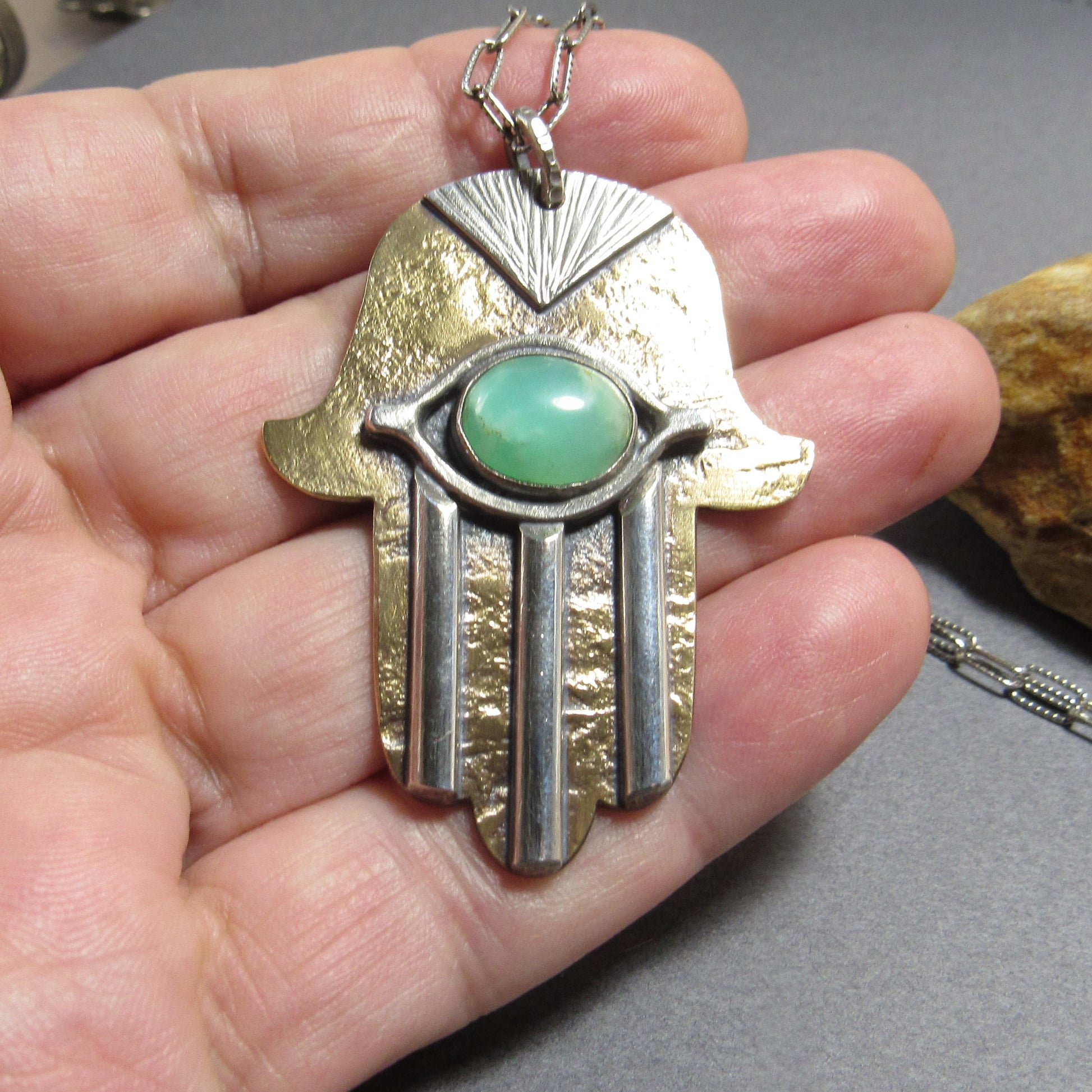 Full view of the Hamsa necklace on a hand, highlighting the detailed sunburst engraving and layered silver “fingers.” Photo background is grey with rock.