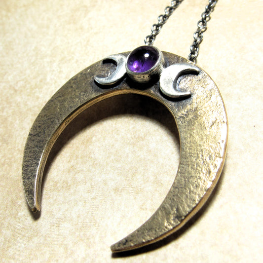 Mixed metal crescent moon pendant featuring an upside-down bronze crescent, a central amethyst cabochon bezel-set in silver, and flanked by two silver crescent moons in a Triple Goddess design. Rustic, worn texture and an oxidized finish.
