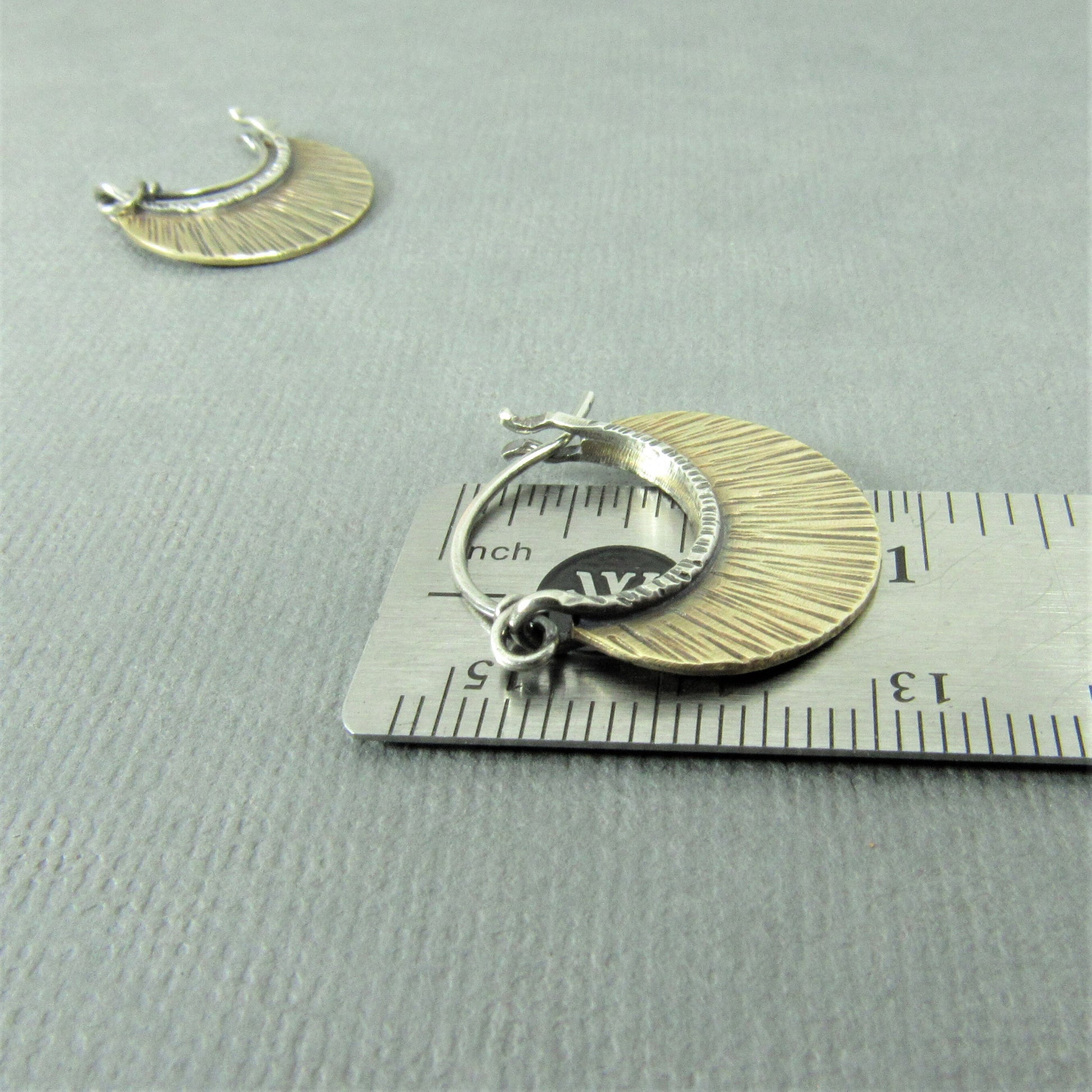Pair of small mixed metal hoop earrings with line texturedesigns on a gray surface with a ruler for scale.