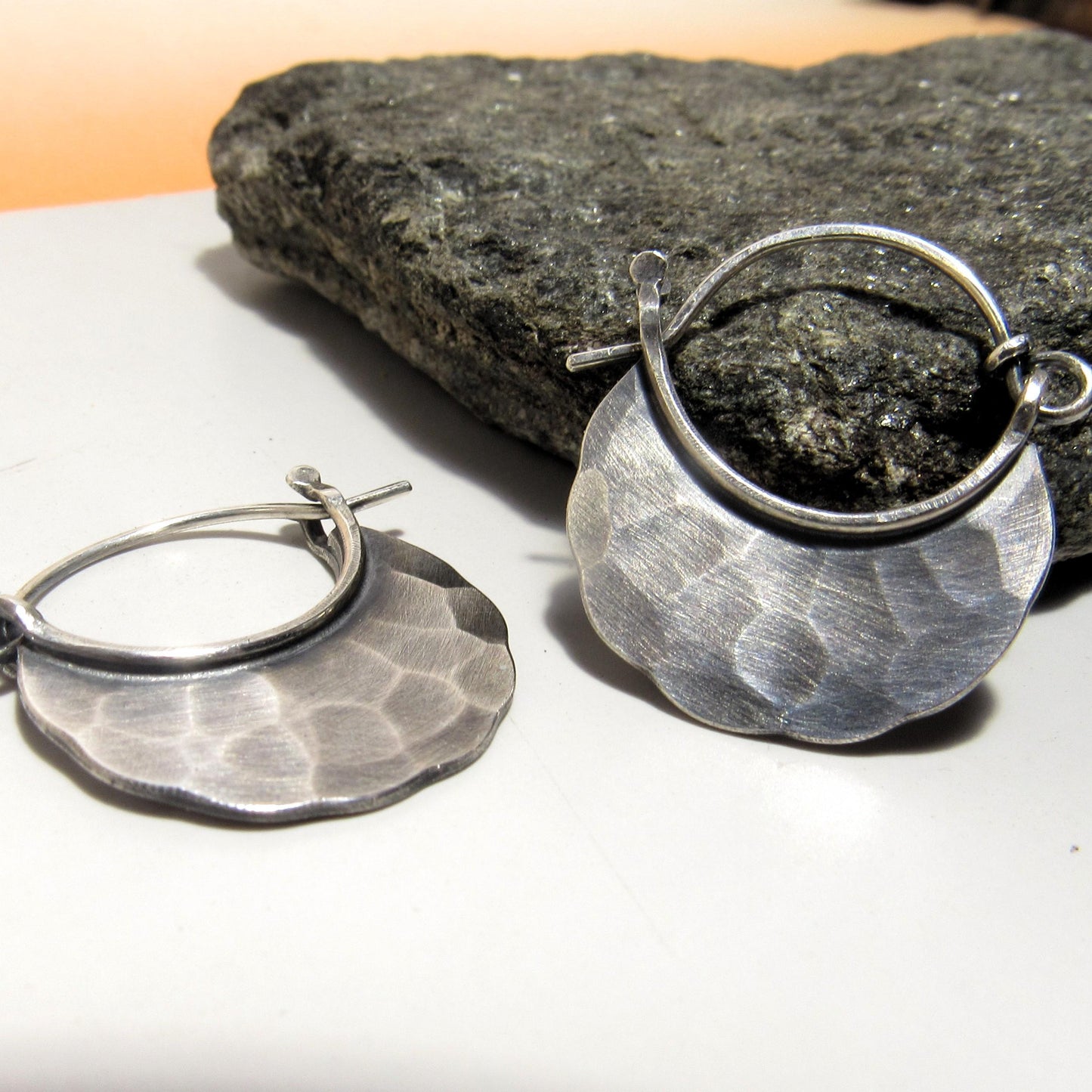 Pair of small silver hoop earrings on a white surface with a rock in the background.