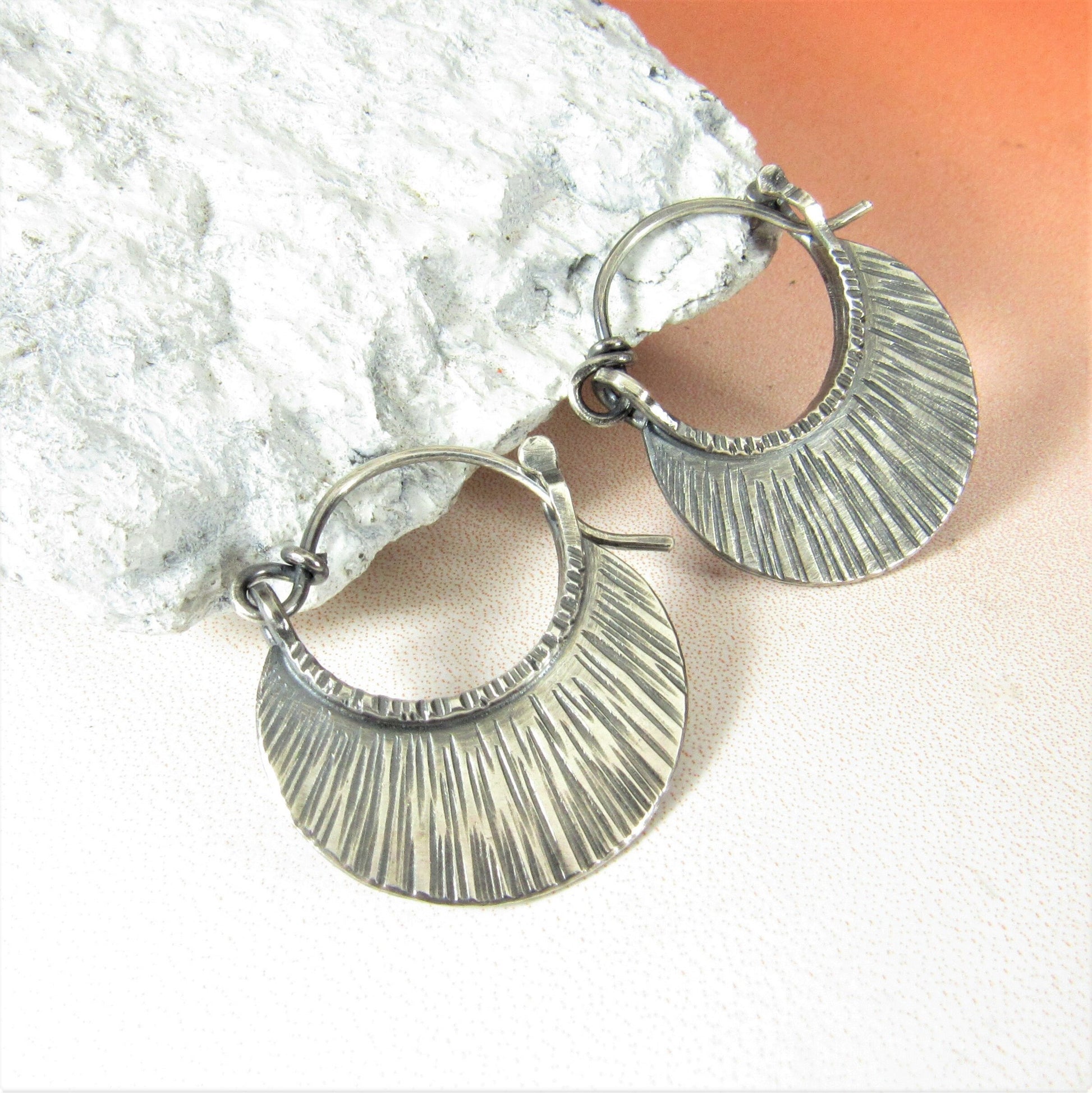 Small textured argentium silver hoop earrings against white rock on orange background.