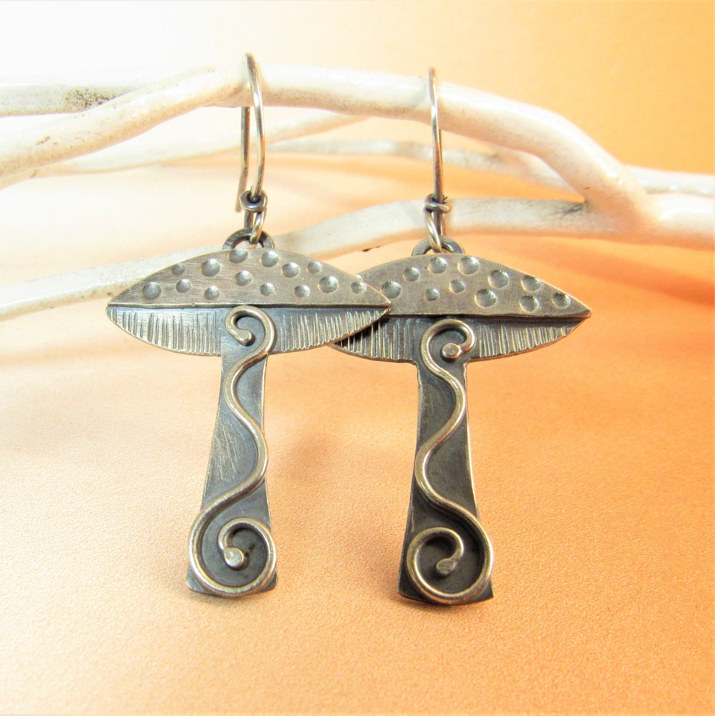 Handcrafted silver mushroom earrings on whte twig and orange background.