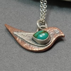 Close up of mixed metal bird pendant by Mocahete. Displayed flat on grey background.