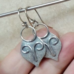 Argentium silver mask earrings with a flourish of wire embellishment. Displayed in hand against neutral background.