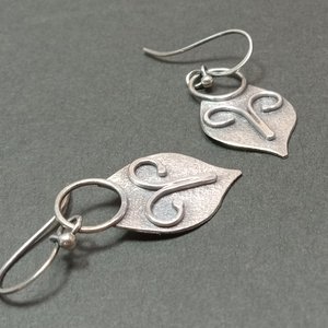 Argentium silver dangle earrings in a mask shape with wire embellishment. Displayed on dark grey background.