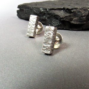 A pair of silver bar stud earrings with a textured finish. The earrings are on a gray surface with dark rock.
