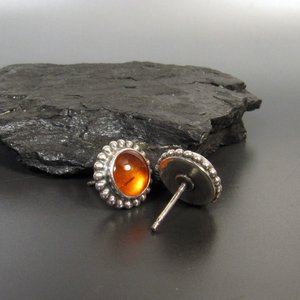A pair of silver stud earrings with a round amber stone in the center. The earrings have a beaded border around the stone, displayed against a black rock on grey background.
