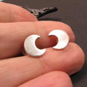 A pair of silver crescent moon earrings. The earrings are small and delicate, and they are perfect for adding a touch of whimsy to any outfit. Photographed in hand.