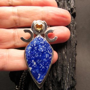 Triple goddess / moon pendant necklace with a faceted citrine and large lapis lazuli cabochon crafted from argentium sterling silver, fine silver. Pendant measures 2 inches from the top and is 7/8 inch across widest part and is suspended on a 18 inch sterling silver chain. Displayed against hand on charred wood.
