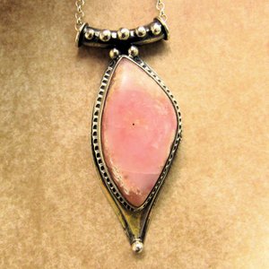 Soft elegance meets artisan craftsmanship in this one of a kind handcrafted pendant featuring a beautifully pastel pink opal set in textured Argentium® sterling silver. The unique, petal-shaped stone in a petal shaped setting radiates gentle warmth in a soothing blush hue—perfect for those who adore subtle statement pieces with soulful charm. 2.25" long x 7/8" wide.
