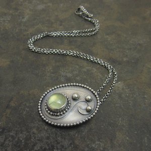 Green prehnite cabochon and argentium sterling silver pendant necklace on dark stone colored background with chain and clasp trailing in the background.