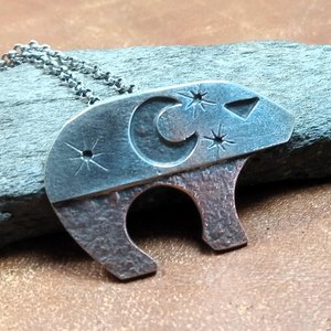 Sterling silver and copper bear pendant necklace inspired by Ursa Major, featuring crescent moon and star cutouts in the silver layer that reveal textured copper beneath, oxidized and polished, on an 18 inch sterling silver chain. Bear measures 3/4" tall x 1.25" long. Displayed against rock and leather.