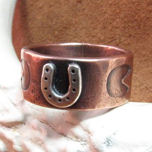 Wide rustically textured copper band with silver horseshoe and horse hoof print stamp motif. Ring has been oxidized and polished giving it a deep and rich patina, and is photographed on grey background propped on a deer antler. Band is made in whole and half sizes from 6-16.