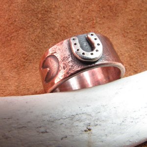 Wide rustically hand textured copper band with silver horseshoe and horse hoof print stamp motif. Ring has been oxidized and polished giving it a deep and rich patina, and is photographed on grey background propped on a deer antler. Band is made in whole and half sizes from 6-16.