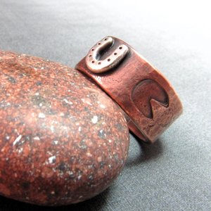 Wide rustically textured copper band with silver horseshoe and horse hoof print stamp motif. Ring has been oxidized and polished giving it a deep and rich patina, and is photographed on grey background propped on a red rock. Band is made in whole and half sizes from 6-16.