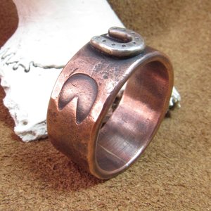 Wide rustically textured copper band with silver horseshoe and horse hoof print stamp motif. Ring has been oxidized and polished giving it a deep and rich patina, and is photographed on grey background stood up in front of a deer antler. Band is made in whole and half sizes from 6-16.