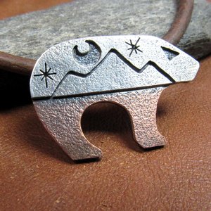 Bear pendant with the top half sculpted in silver with moon and star cutouts and the lower body in richly textured copper, joined harmoniously through traditional metalsmith techniques. The pendant is suspended on a sturdy 18-22 inch leather cord finished with our handmade Argentium silver findings for durability and comfort.Bear measures 1.25" long and is .75" tall. Displayed on brown leather against rock.