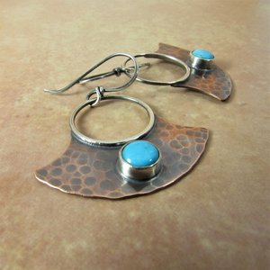 May include: A pair of handmade earrings with a hammered copper fan shape and a turquoise stone set in silver. The earrings have a silver hoop and a silver hook.