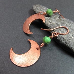 Side view of Handmade copper crescent moon dangle earrings with faceted green chrysoprase beads. Earrings measure 2 and 1/8" long and the moon is 3/8" wide at widest point.The ear wires are solid, pure copper. Displayed on dark background against rock.