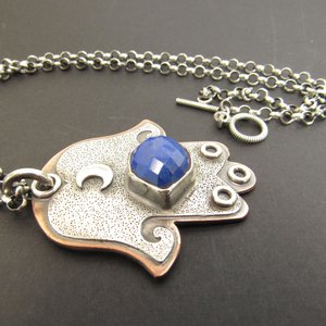 Mixed metal, copper and argentium sterling silver hamsa pendant necklace with lapis lazuli cabochon and crescent moon detail. 19 inch ss chain and findings. Hamsa is 1.5" wide x 1.75" tall. Displayed against dark background.