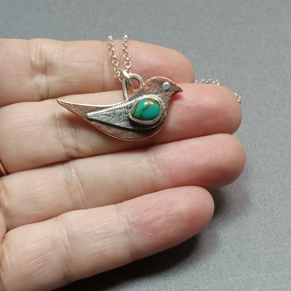 Copper, silver and turquoise bird pendant necklace by Mocahete. Displayed in hand.