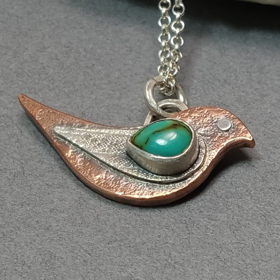 Close up of mixed metal bird pendant by Mocahete. Displayed flat on grey background.
