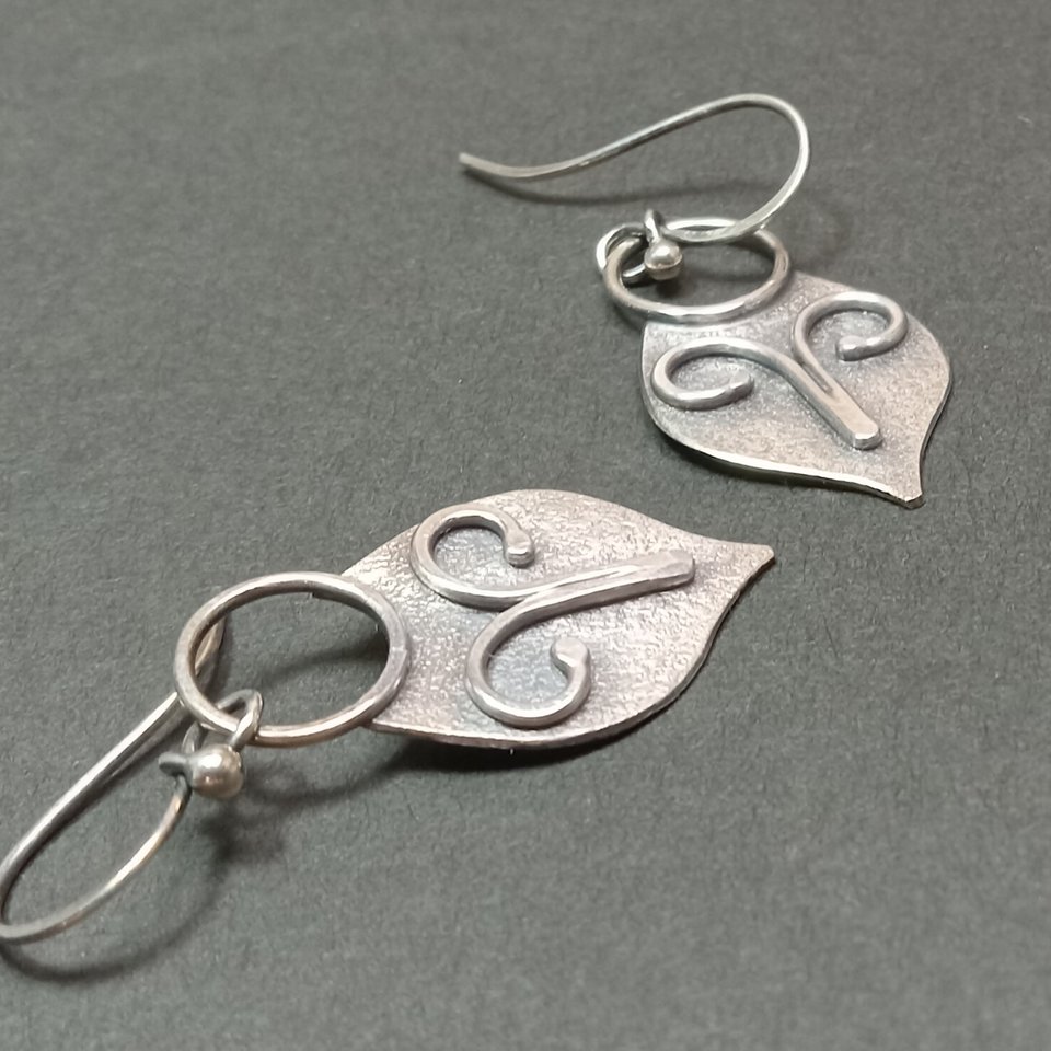 Argentium silver dangle earrings in a mask shape with wire embellishment. Displayed on dark grey background.
